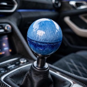 May include: A spherical gear shift knob featuring a glossy blue finish with a swirling white pattern in the upper half and a dark blue glitter effect in the lower half, mounted on a black metal stem and a black leather shift boot in a vehicle.