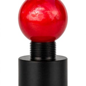 May include: A gear shift knob featuring a smooth, glossy red spherical top attached to a black cylindrical base with a threaded metal neck. The design consists of a vibrant red ball mounted on a matte black metal component with visible threading.