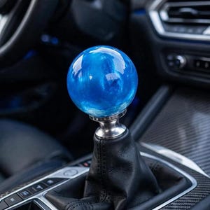 May include: A spherical blue gear shift knob with a swirling, translucent finish, mounted on a silver-toned metal base and a black leather boot. The gear shifter is positioned within a vehicle center console featuring carbon fiber trim accents.