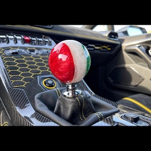 May include: A spherical gear shift knob featuring a glossy finish with vertical stripes in red, white, and green. The knob is mounted on a chrome-finished base attached to a black leather gear shift boot with yellow stitching inside a vehicle interior.