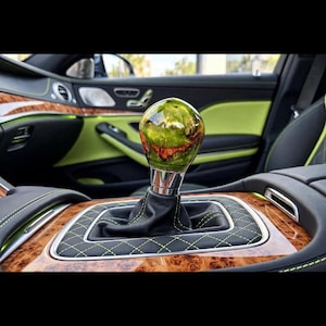 Può includere: Un pomello del cambio sferico con finitura in resina lucida verde e marrone e base metallica, montato su una cuffia in pelle nera con cuciture a rombi verde lime, inserito in una console centrale con finiture in legno.