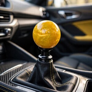 Peut inclure: Un pommeau de levier de vitesse sphérique jaune avec une finition marbrée brillante fixé sur une base en métal noir. Le pommeau repose sur un soufflet en cuir noir avec des coutures jaunes, installé dans une console centrale avec garniture carbone.