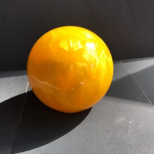 May include: A spherical, glossy, yellow-orange object. The surface has a subtle, swirling pattern, and the object appears to be made of a translucent material. The sphere is resting on a dark grey surface, with a shadow cast to the side.