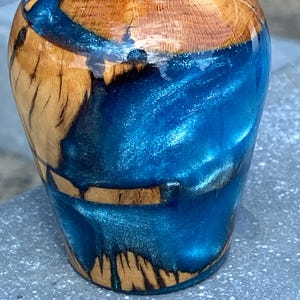 Puede incluir: Un objeto cilíndrico elaborado con madera natural de color marrón claro y resina azul vibrante y brillante. La madera presenta vetas visibles, mientras que la resina azul brillante fluye por el centro en un diseño arremolinado, metálico y translúcido.
