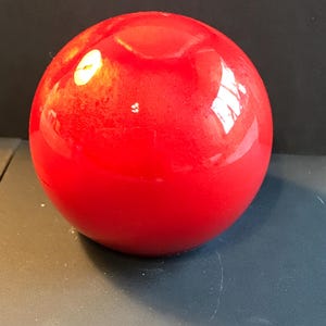 Puede incluir: Una esfera roja brillante y sólida, que refleja la luz. La superficie lisa de la bola es el punto focal, con una ligera hendidura en la parte superior. El objeto es probablemente un elemento decorativo o parte de un conjunto más grande.