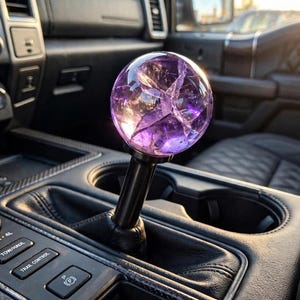 May include: A spherical purple gear shift knob with internal crackled detailing mounted on a black cylindrical shaft, positioned within the centre console of a vehicle featuring black leather upholstery and various control buttons and dials.