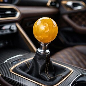 May include: A spherical gear shift knob with a translucent, marbled amber and gold finish, mounted on a silver-toned metal base. The knob is attached to a black leather shift boot featuring visible gold stitching, set within a carbon fiber console.