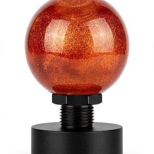 May include: A spherical gear shift knob featuring a translucent orange resin finish with embedded gold glitter particles. The knob is attached to a black metal threaded adapter and a cylindrical black base, creating a contrast in texture and color.