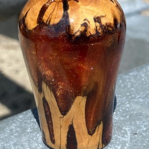 May include: A cylindrical object crafted from a combination of natural light-colored wood and a deep, translucent amber resin infused with shimmering copper-toned glitter. The wood features prominent grain patterns that extend upward into the resin.