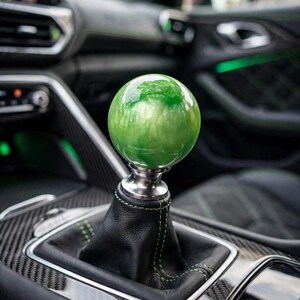 May include: A spherical gear shift knob with a glossy, marbled green and white finish. The knob is mounted on a silver-toned metal base, which connects to a black leather shift boot featuring decorative green stitching details.