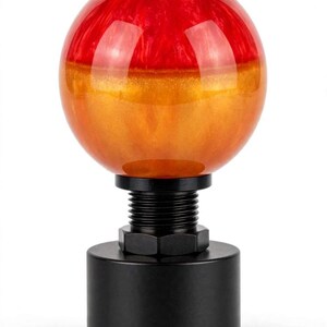 May include: A spherical gear shift knob featuring a two-tone design with a translucent red top half and a shimmering orange bottom half. The knob is attached to a black cylindrical metal base with a threaded neck and a hexagonal locking nut.