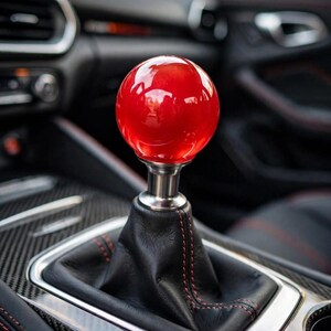Peut inclure: Un pommeau de levier de vitesse sphérique rouge brillant monté sur une base en argent métallique. Le pommeau est fixé à un soufflet en cuir noir avec des coutures rouges, situé dans la console centrale d'un véhicule avec des garnitures en carbone.