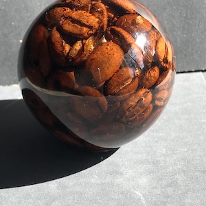 May include: A transparent, spherical paperweight containing many roasted coffee beans. The beans are a deep brown, and the sphere is clear, allowing light to pass through and highlight the beans' texture. The paperweight rests on a grey surface.