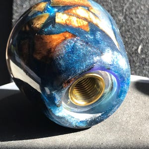May include: A decorative, handcrafted knob with a blue and gold resin design. The knob features a mix of blue resin with embedded wood pieces and a brass threaded insert for attachment. The overall shape is rounded and smooth.
