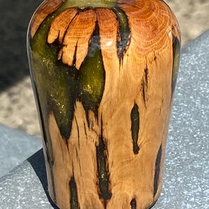 Puede incluir: Un objeto cilíndrico fabricado en madera natural de color marrón claro con un veteado prominente, que presenta fisuras verticales y huecos rellenos de una resina verde oliva translúcida y brillante, creando un acabado exterior liso y pulido.