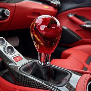 May include: A spherical gear shift knob featuring a glossy red and dark wood grain resin finish, mounted on a chrome base with a black leather boot, positioned within a vehicle interior characterised by red leather upholstery and carbon fibre trim.