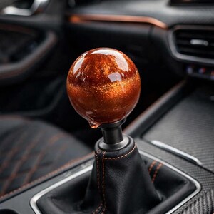 May include: A spherical gear shift knob featuring a glossy, translucent amber-colored resin finish with embedded gold glitter particles. The knob is mounted on a black metal stem attached to a black leather shift boot with orange stitching.