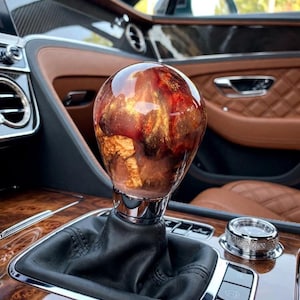 May include: A spherical gear shift knob featuring a polished resin finish with swirling patterns of deep red, amber, and metallic gold leaf accents. The knob is mounted on a black leather boot with a chrome base, situated in a luxury vehicle interior.