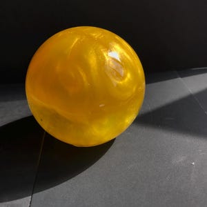 May include: A golden, spherical object with a swirling, textured appearance. The sphere is illuminated, creating highlights and shadows. The object is set against a dark background, emphasizing its color and form. The overall theme is decorative.