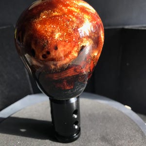 May include: A custom shift knob with a glossy, spherical top. The top is a mix of red, brown, and gold, with a swirling, marbled effect. The base is black and cylindrical with drilled holes. The knob is designed for a vehicle's gear shift.