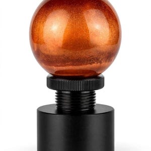 Könnte beinhalten: Ein kugelförmiger Schaltknauf mit glänzender, metallisch kupferfarbener Oberfläche und feinen Glitzerpartikeln. Die Kugel ist auf einem schwarzen zylindrischen Sockel mit einem Gewindeanschluss und einem geriffelten Einstellring montiert.