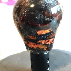 May include: A black and clear gear shift knob with a wood grain design. The top of the knob is a glossy black with silver glitter. The wood grain is a mix of black and orange.