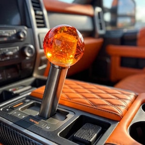 May include: A spherical gear shift knob featuring a translucent orange resin top with internal bubble details, mounted on a cylindrical brushed metal shaft. The gear shifter is positioned in the center console of a vehicle with orange upholstery.
