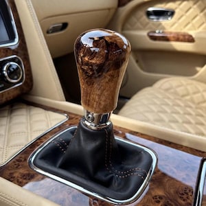 May include: A gear shift knob featuring a polished, swirling brown burl wood grain finish, mounted on a black leather boot with visible stitching, positioned within a vehicle interior with tan quilted upholstery and wood trim accents.