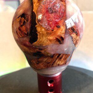 Puede incluir: Un pomo de cambio personalizado con un diseño único. El pomo presenta una combinación de madera y resina, con una combinación de colores rojo y marrón. La base es roja con dos agujeros. La forma general es redonda.