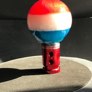 May include: A spherical gear shift knob featuring horizontal bands of red, white, and blue resin with a glossy finish. The knob is attached to a cylindrical red metal base that includes two circular side holes and a threaded bottom section.