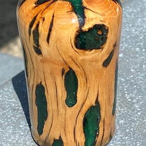 May include: A cylindrical object crafted from light brown wood with a polished finish, featuring irregular natural grain patterns and voids filled with a smooth, opaque dark green resin that creates a striking contrast against the wood texture.