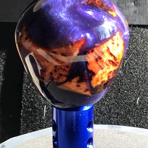 May include: A custom shift knob with a blue base and a spherical top. The top is a mix of deep purple and orange hues, with a glossy finish. The base is a vibrant blue with several drilled holes.