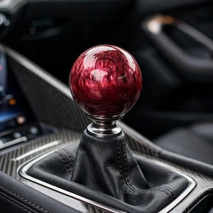 Può includere: Un pomello del cambio sferico con un motivo marmorizzato lucido rosso scuro e nero. Il pomello è montato su una base in argento metallizzato, collegata a una cuffia in pelle nera. L'insieme è inserito in una console centrale in carbonio.