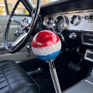 May include: A spherical gear shift knob featuring a horizontal tri-color design with a glossy red top section, a white middle section with swirling textures, and a blue bottom section, mounted on a ribbed chrome metal shaft inside a vehicle.