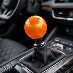 May include: A spherical orange gear shift knob with a glossy finish, mounted on a black leather shift boot featuring orange stitching. The gear shifter is positioned within a vehicle center console that includes carbon fiber trim and buttons.