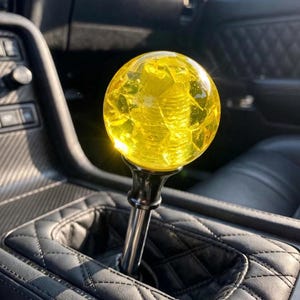 May include: A spherical yellow gear shift knob with a cracked glass-like texture, mounted on a metallic silver shaft with a black base, positioned within a vehicle interior featuring black quilted leather upholstery and dark trim components.