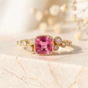 Könnte beinhalten: Ein Goldring mit einem zentralen, kissenförmigen rosa Edelstein, der von einer Gruppe kleinerer, rund geschliffener weißer Steine umgeben ist. Die Ringschiene besteht aus poliertem gelbem Metall und die Fassung weist feine Milgrain-Details auf.