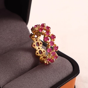 Könnte beinhalten: Ein goldener Ring mit einer doppelten Reihe kreisförmiger, leuchtend pinker Edelsteine in einem durchbrochenen, gewellten Metalldesign. Der Ring besteht aus einem durchgehenden Muster miteinander verbundener Goldglieder, die jeden Stein halten.