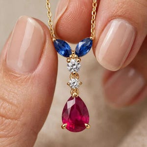 Può includere: Una collana in oro con un pendente caratterizzato da una pietra rossa a forma di pera in basso, due piccole pietre chiare rotonde sopra e due pietre blu taglio marquise in alto, montate su una sottile catena d'oro.