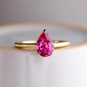 Può includere: Un anello in oro con una singola gemma rosa vibrante, sfaccettata e a forma di pera, montata su una struttura a quattro griffe. Il gambo dell'anello ha una finitura dorata opaca e liscia, che contrasta con la superficie brillante e riflettente della pietra.