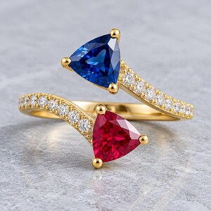 May include: A gold bypass ring featuring two trillion-cut gemstones, one deep blue and one vibrant red, set at the open ends. The band is accented with a row of small, clear, round-cut stones along each side, creating a sparkling, symmetrical design.