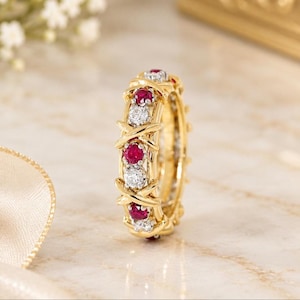 Puede incluir: Un anillo de oro amarillo con un patrón alterno de rubíes rojos redondos y diamantes blancos transparentes. Las piedras preciosas están engastadas en una serie de motivos de cruz en forma de X que rodean toda la circunferencia del anillo.