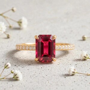 Könnte beinhalten: Ein Goldring mit einem großen, rechteckigen, smaragdgeschliffenen tiefroten Edelstein in einer Vier-Krappen-Fassung. Die Ringschiene besteht aus Gelbgold und ist mit einer Reihe kleiner, runder, klarer weißer Diamanten besetzt.