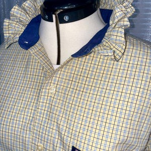 Könnte beinhalten: Ein gelb-blau kariertes Hemd mit Rüschenkragen und blauem Besatz. Das Hemd hat eine Knopfleiste und eine kleine Tasche mit dem "CHAPS"-Logo.