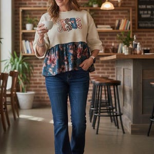 May include: A cream-colored long-sleeve top with a peplum hem featuring a dark blue floral pattern and the word JESUS printed in a multi-patterned font across the chest, paired with dark blue denim jeans and patterned slip-on sneakers.