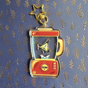 May include: A clear acrylic blender keychain with red and yellow accents. The blender has a handle and the word "BLEND" on the front. A gold star-shaped charm and a gold clasp are attached to the top.