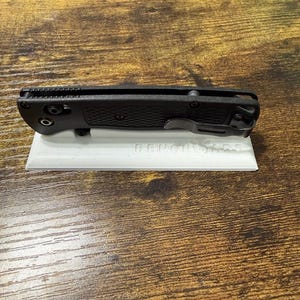 Puede incluir: Una navaja plegable negra con un mango texturizado y un clip para el bolsillo, apoyada sobre una superficie blanca. La navaja está cerrada, mostrando su perfil lateral. El nombre de la marca es visible en la superficie blanca.