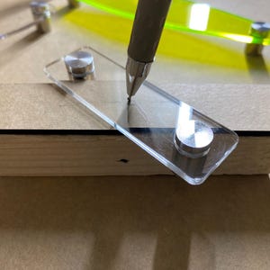 Könnte beinhalten: Eine rechteckige, klare Acrylplatte mit zwei runden, silberfarbenen Metallabstandshaltern, die auf einer flachen braunen Oberfläche liegt. Die Spitze eines grauen Stifts berührt die Mitte der Acrylplatte, die auf einem hellen Holzblock ruht.