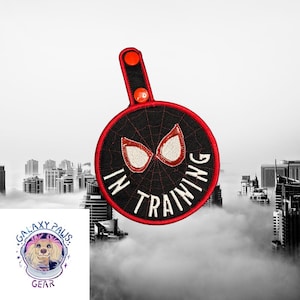 Puede incluir: Un parche negro y rojo con las palabras "IN TRAINING" y un diseño de ojos de araña. El parche tiene un borde rojo y un asa roja con dos botones rojos. El fondo es un paisaje urbano.