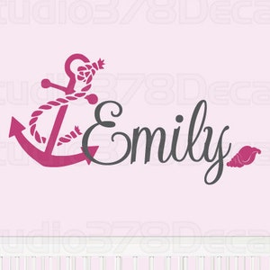 Puede incluir: Ancla rosa con un diseño de cuerda y el nombre Emily en letra gris oscura. Una pequeña concha rosa está a la derecha del nombre. El diseño se muestra en una cuna blanca.